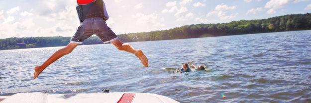 Kids jumping off a boat into the lake. Focus on boys legs and boat, lens flares from sun.