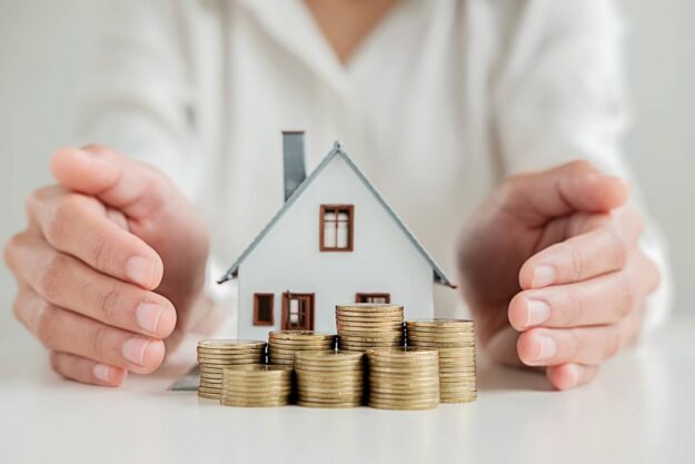 homeowners-insurance Hands protecting a small house model surrounded by stacks of coins, symbolizing home finance and investment.